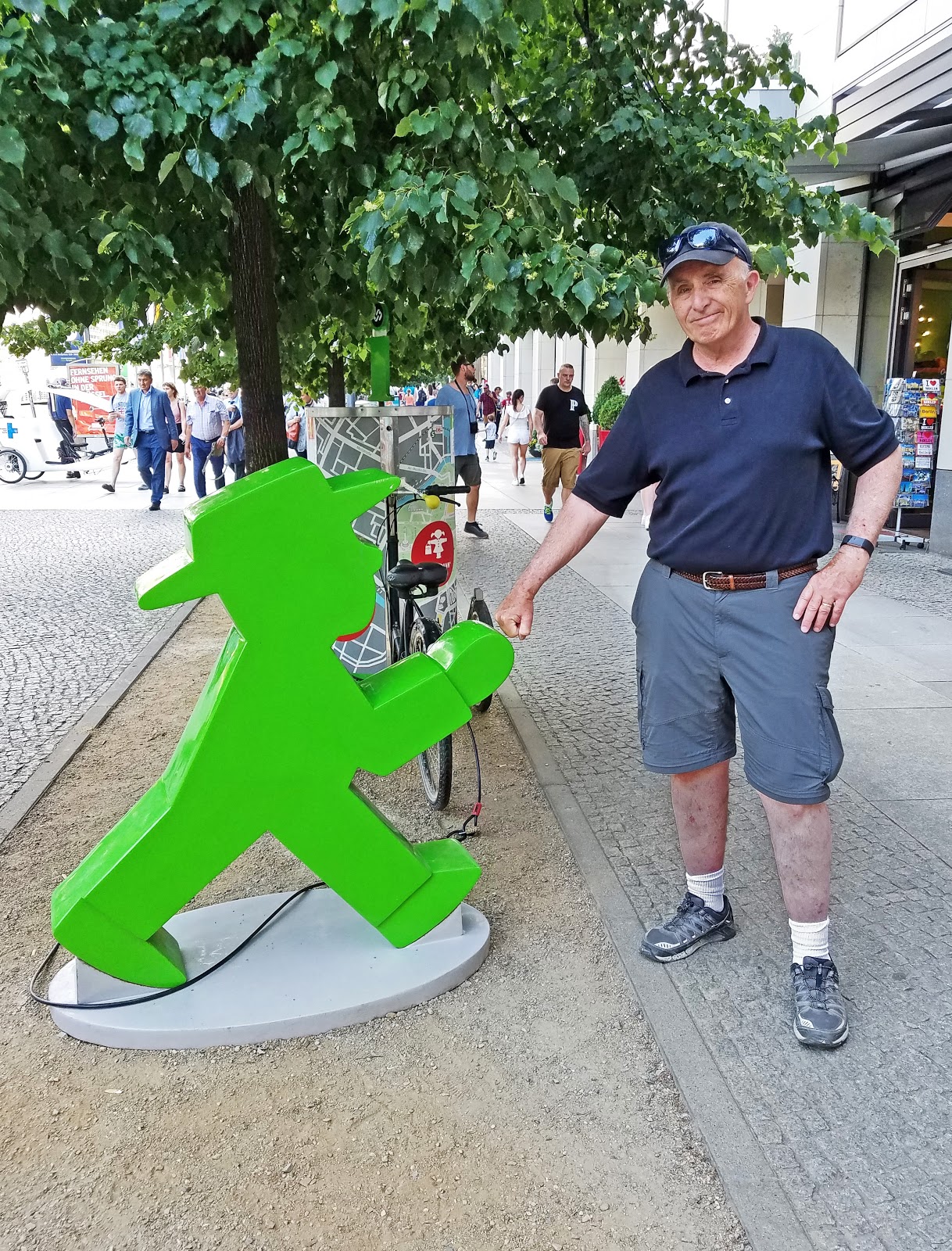 Figurine publicitaire Ampelmann en extérieur 1 Mężczyzna stojący obok dużej zielonej figury przedstawiającej chodzącego człowieka na chodniku, w tle drzewa i ludzie.