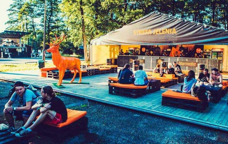 Cerf Jägermeister 1 Ludzie siedzący na pomarańczowych poduszkach na drewnianej platformie przed namiotem z napisem "STREFA JELENIA".