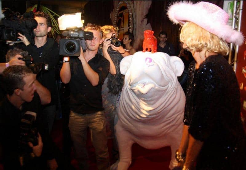Grande figure de bouledogue 2 Kobieta w różowym kapeluszu pozuje na czerwonym dywanie obok dużej, białej rzeźby psa, otoczona przez fotoreporterów.