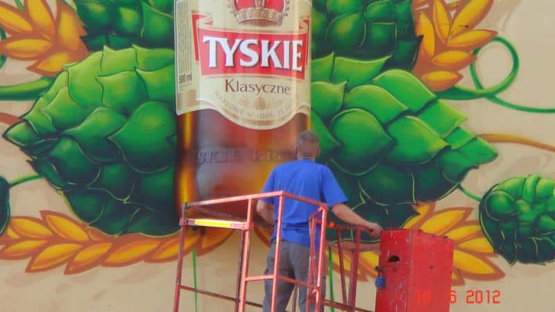 Fresque tridimensionnelle TYSKIE 4 Mężczyzna na podnośniku malujący mural z butelką piwa Tyskie Klasyczne i chmielami.