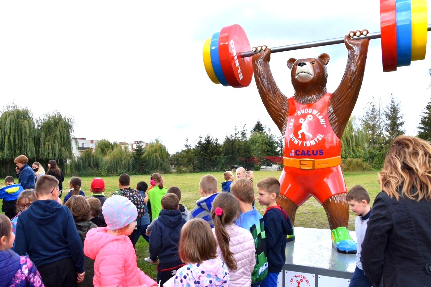 Grand Ours - Championnats d'Haltérophilie 4 Dzieci z nauczycielkami oglądają dużą figurę misia podnoszącego sztangę na zewnątrz.