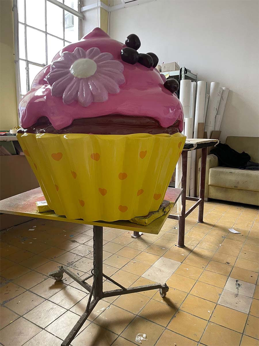 Un cupcake coloré pour la pâtisserie Sweet Home à Lagos, au Portugal 2 Duża, kolorowa rzeźba muffinki w żółtej papilotce z serduszkami, z różowym lukrem, ozdobiona kwiatkiem i wiśniami, umieszczona na stole w pracowni.