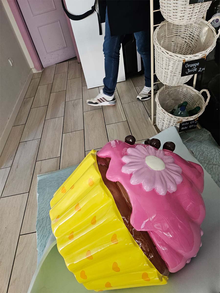Un cupcake coloré pour la pâtisserie Sweet Home à Lagos, au Portugal 3 Rzeźba wielkiego babeczki w pastelowych kolorach stojąca na podłodze w kawiarni obok osoby w dżinsach i białych butach.