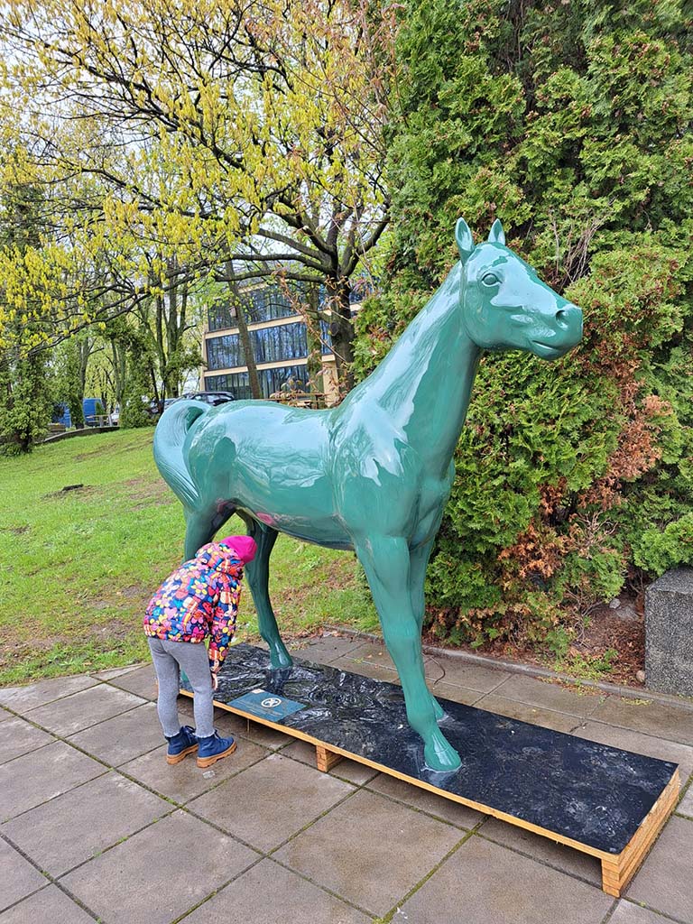 Les sculptures colorées de chevaux sur la piste de Partynice à Varsovie 4 Dziecko w kolorowej kurtce zaglądające pod rzeźbę zielonego konia na zewnątrz w parku.