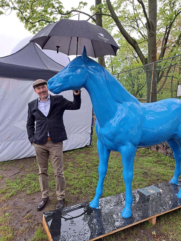 Les sculptures colorées de chevaux sur la piste de Partynice à Varsovie 2 Mężczyzna trzymający parasol nad niebieską statuetką konia.