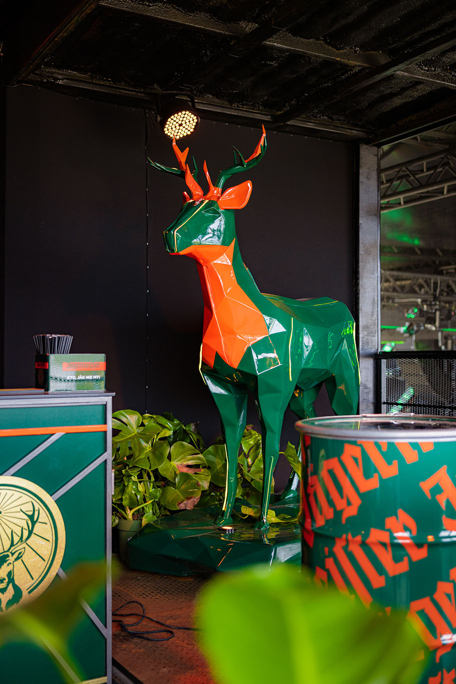 Cerfs Jägermeister avec Éclairage LED 2 Pomarańczowo-zielona rzeźba jelenia stojąca w otoczeniu roślin i zielono-pomarańczowych elementów dekoracyjnych.
