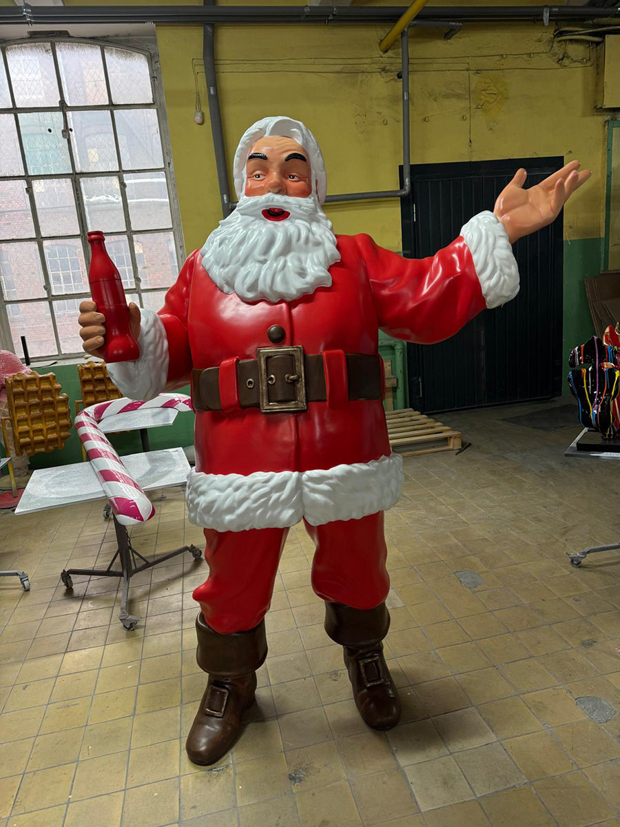 Tout le monde peut être Père Noël : la campagne de Noël de Coca-Cola avec des figurines de Père Noël 17 Figurka Świętego Mikołaja w czerwonym stroju i brązowych butach stojąca na podłodze w pomieszczeniu z żółto-zielonymi ścianami i dużym oknem, trzymająca czerwoną butelkę w jednej ręce i gestykulująca drugą.