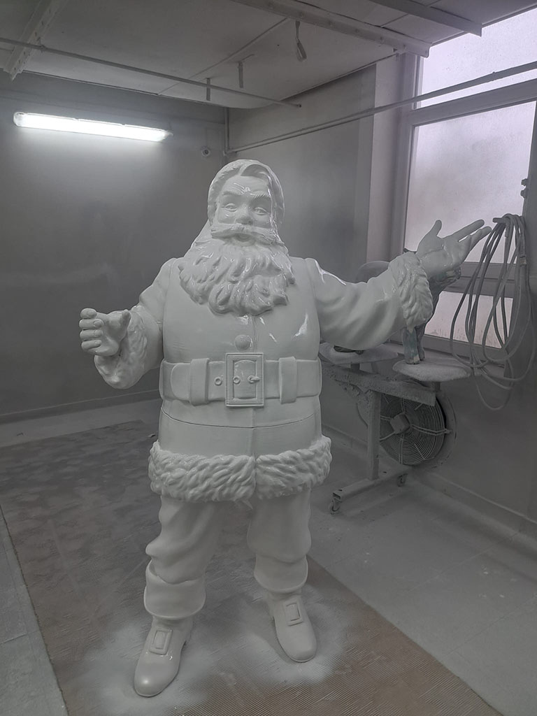 Tout le monde peut être Père Noël : la campagne de Noël de Coca-Cola avec des figurines de Père Noël 18 Biały, niepomalowany posąg Świętego Mikołaja w pracowni malarskiej, z uniesionymi rękoma, obok okno i wentylator.