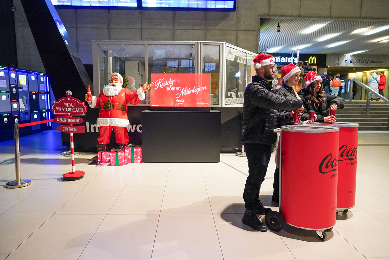 Tout le monde peut être Père Noël : la campagne de Noël de Coca-Cola avec des figurines de Père Noël 3 Trzy osoby w czapkach św. Mikołaja stoją przy czerwonych pojemnikach Coca-Coli w hali dworca, obok stoi figura św. Mikołaja z butelką Coca-Coli i znakiem "Witaj w Katowicach" oraz cyfrowym ekranem z napisem reklamowym.