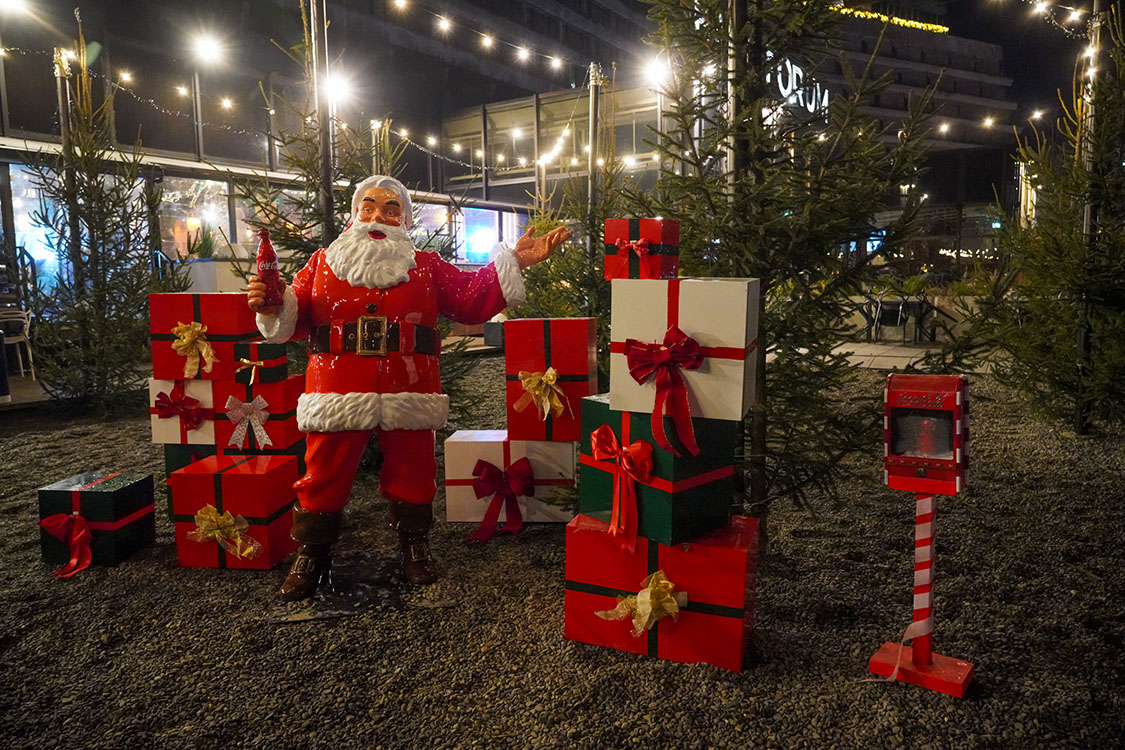 Tout le monde peut être Père Noël : la campagne de Noël de Coca-Cola avec des figurines de Père Noël 1 Figurka Świętego Mikołaja trzymającego butelkę Coca-Coli, otoczona licznymi prezentami w czerwonych, białych i zielonych opakowaniach z kokardami, ustawiona na żwirowym podłożu na tle drzewek iglastych i oświetlenia świątecznego nocą