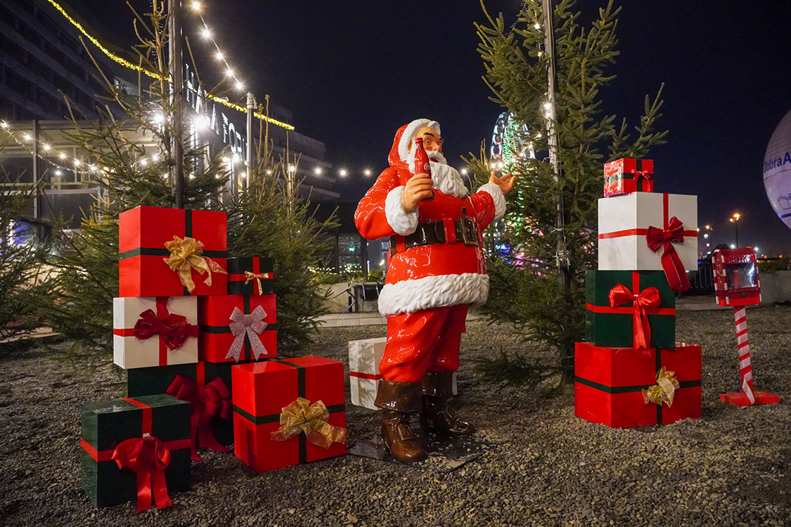 Tout le monde peut être Père Noël : la campagne de Noël de Coca-Cola avec des figurines de Père Noël 2 Figurka Mikołaja trzymającego butelkę napoju w otoczeniu świątecznie udekorowanych prezentów i małych drzewek iglastych oświetlonych lampkami nocą w plenerze.
