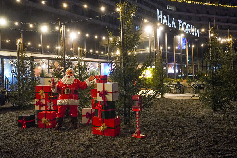 Tout le monde peut être Père Noël : la campagne de Noël de Coca-Cola avec des figurines de Père Noël 4 Rankiem na zewnątrz hali Forum ozdobionej światełkami, stoi duża figurka Świętego Mikołaja otoczona wysokimi choinkami i kolorowymi, ozdobnymi prezentami; w tle szklana fasada i neonowa nazwa Hala Forum.