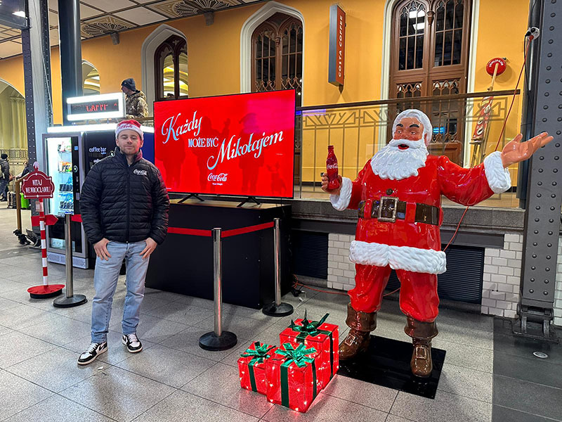 Tout le monde peut être Père Noël : la campagne de Noël de Coca-Cola avec des figurines de Père Noël 5 Mężczyzna w czapce Mikołaja stoi obok dużej figurki Świętego Mikołaja trzymającego butelkę Coca-Coli, obok znajdują się podświetlane prezenty, a za nimi czerwony ekran z napisem "Każdy może być Mikołajem" w dworcu kolejowym lub podobnym wnętrzu.