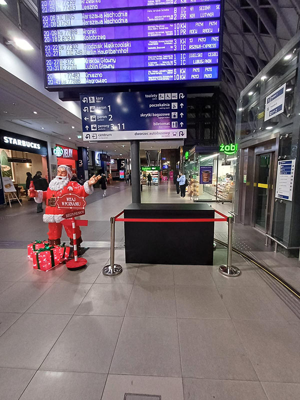 Tout le monde peut être Père Noël : la campagne de Noël de Coca-Cola avec des figurines de Père Noël 7 Wnętrze dworca z tablicą odjazdów pociągów na górze, na dole figura Mikołaja z prezentami i napisem Witaj w Poznaniu oraz sklepami Starbucks i Żabka w tle.