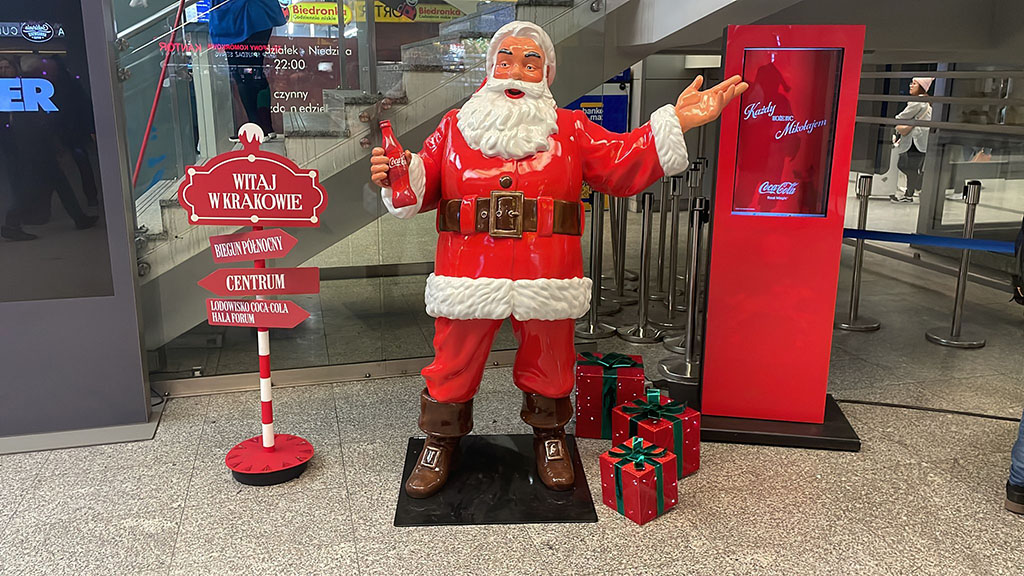 Tout le monde peut être Père Noël : la campagne de Noël de Coca-Cola avec des figurines de Père Noël 8 Figurka Mikołaja w czerwonym stroju trzymającego butelkę Coca-Coli, stojąca obok czerwonego stojaka z ekranem oraz tabliczki z napisem Witaj w Krakowie i kierunkami do Bieguna Północnego, centrum i lodowiska Coca-Coli, a także przedmiotów wyglądających jak prezenty na podłodze, w nowoczesnym pomieszczeniu z szarym posadzką i przeszklonym wejściem w tle
