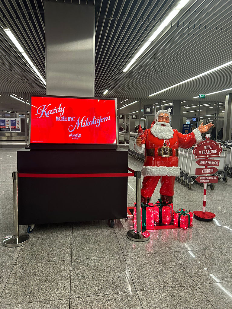 Tout le monde peut être Père Noël : la campagne de Noël de Coca-Cola avec des figurines de Père Noël 9 Figurka Świętego Mikołaja z butelką Coca-Coli i dekoracyjnymi prezentami przy informacji lotniskowej, obok stoi czerwony ekran z napisem "Każdy może być Mikołajem" oraz tablica powitalna z napisem "Witaj w Krakowie" w nowoczesnym terminalu lotniska.