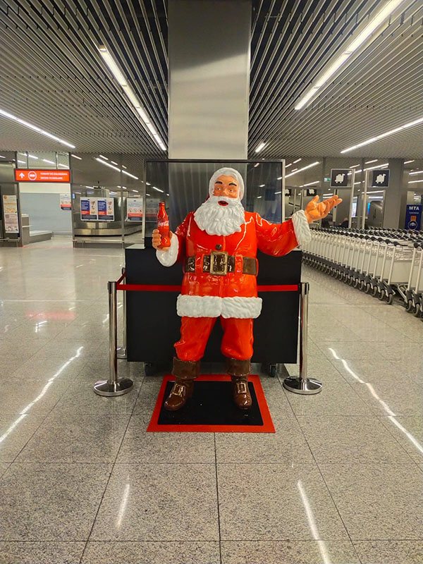 Tout le monde peut être Père Noël : la campagne de Noël de Coca-Cola avec des figurines de Père Noël 10 Figurka Mikołaja w czerwonym stroju z białym futrem stojąca na czarno-czerwonym podłożu, trzymająca czerwoną butelkę, w nowoczesnym hali z metalowymi sufitami i rzędem wózków po prawej stronie