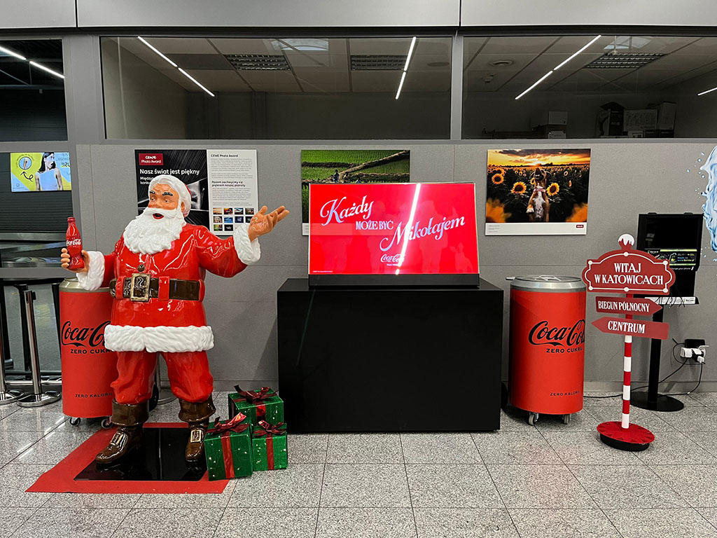 Tout le monde peut être Père Noël : la campagne de Noël de Coca-Cola avec des figurines de Père Noël 12 Figurka świętego Mikołaja w czerwonym ubraniu trzymająca butelkę Coca-Coli, stojąca obok dwóch zielonych prezentów z czerwonymi kokardami, na tle szarej ścianki z plakatami i czerwonych pojemników na napoje Coca-Cola Zero, oraz czerwony znak z napisami witającymi w Katowicach i wskazującymi kierunki.