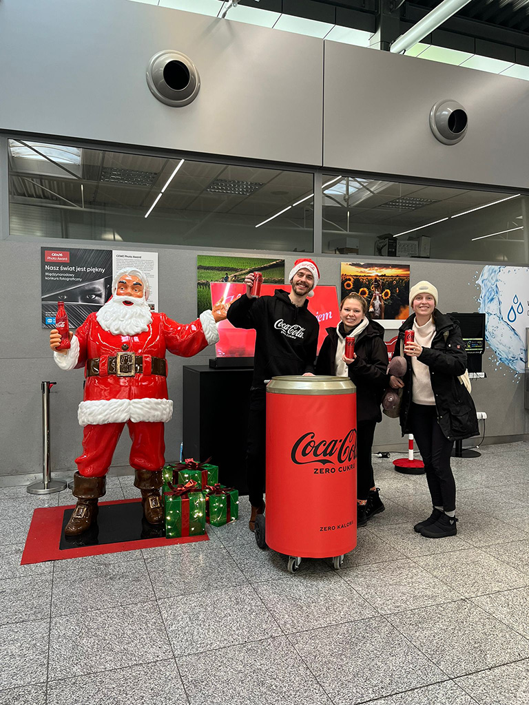 Tout le monde peut être Père Noël : la campagne de Noël de Coca-Cola avec des figurines de Père Noël 13 Trzy osoby stoją obok dużej figury Mikołaja w czerwonym stroju trzymającego butelkę Coca-Coli, każda z nich trzyma puszkę Coca-Coli, obok stoi duża puszka Coca-Coli na kółkach, tło to wnętrze budynku z szarymi ścianami i gablotami ze zdjęciami.