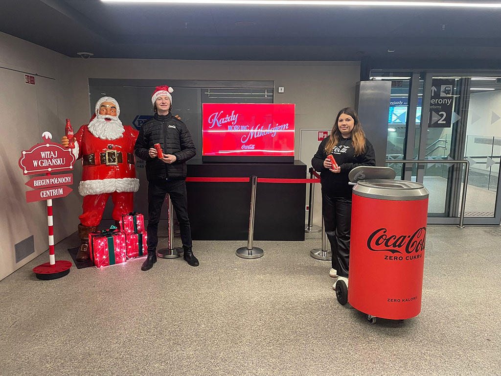 Tout le monde peut être Père Noël : la campagne de Noël de Coca-Cola avec des figurines de Père Noël 14 Dwoje ludzi trzymających puszki Coca-Coli stoi obok dużej czerwonej figurki Mikołaja i czerwonego śmietnika w kształcie puszki Coca-Coli w jasnym, nowoczesnym wnętrzu na tle ekranu z napisem w języku polskim i czerwonym stojakiem z napisem powitalnym.