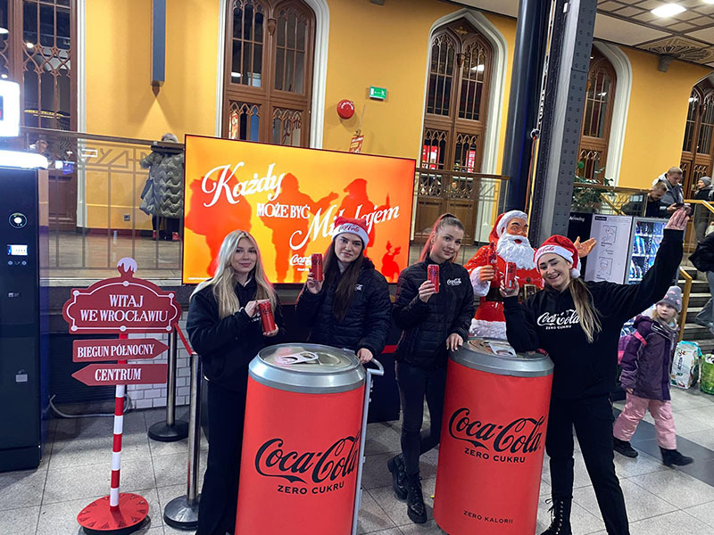Tout le monde peut être Père Noël : la campagne de Noël de Coca-Cola avec des figurines de Père Noël 16 Cztery młode kobiety w ciemnych kurtkach i czapkach Mikołaja pozują z puszkami Coca-Coli przy dwóch dużych stojakach w kształcie puszek Coca-Coli Zero Cukru, na tle ekranu z napisem "Każdy może być Mikołajem" i figury Świętego Mikołaja.